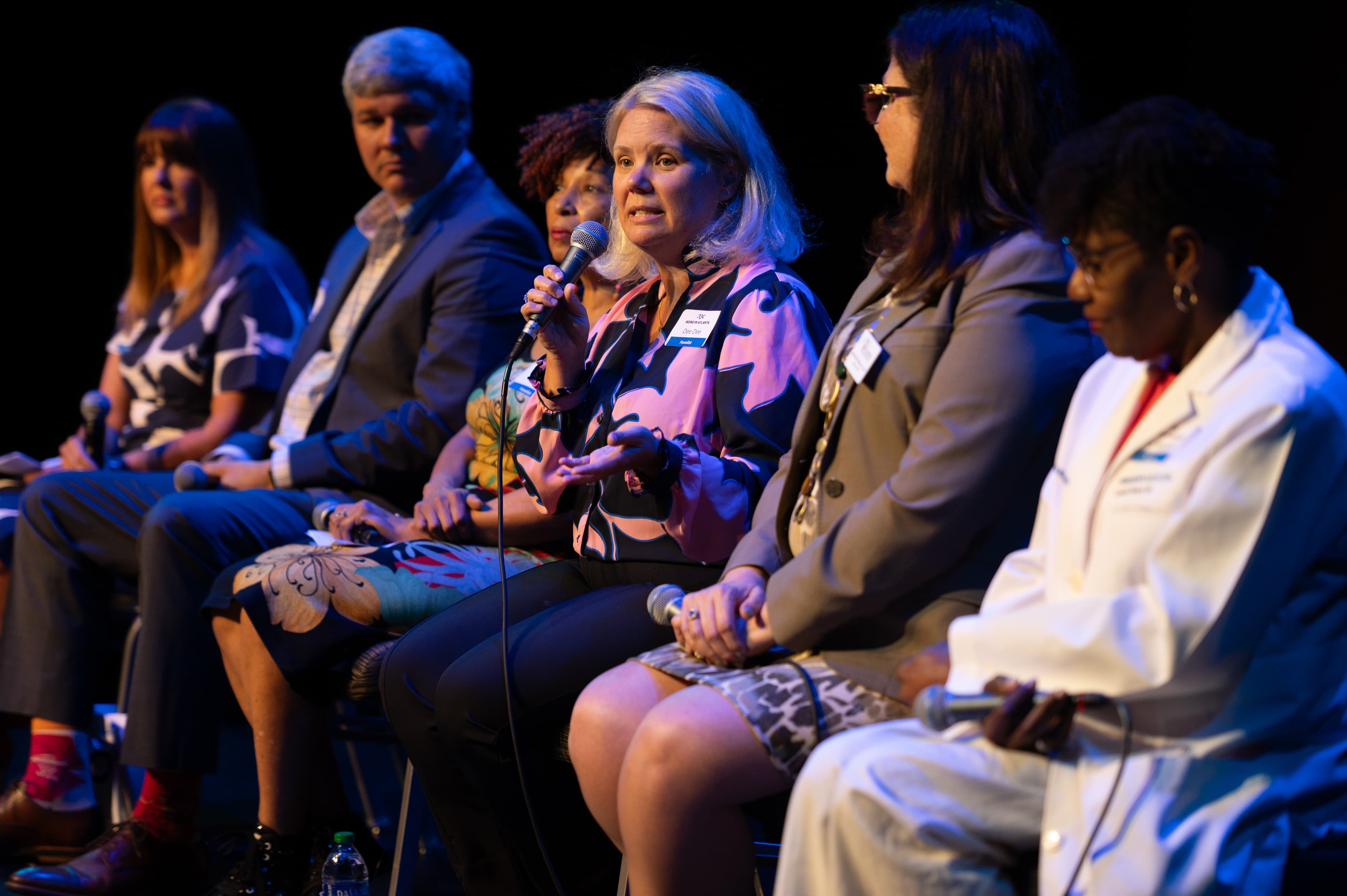 Event coverage photograph of a professional panel discussion with six business panelists seated on a darkened stage with dramatic blue and pink lighting