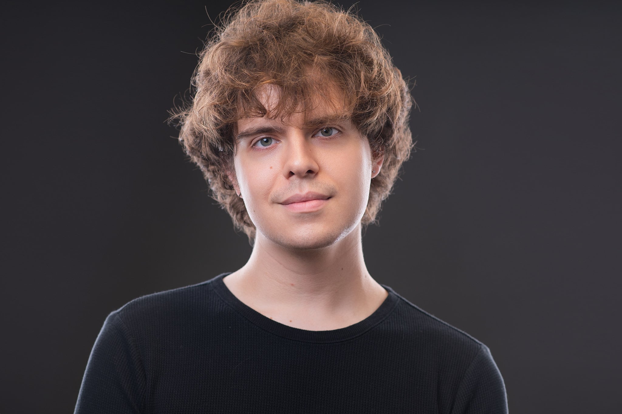 Artistic young male actor headshot with shoulder-length wavy hair and serious expression against grey