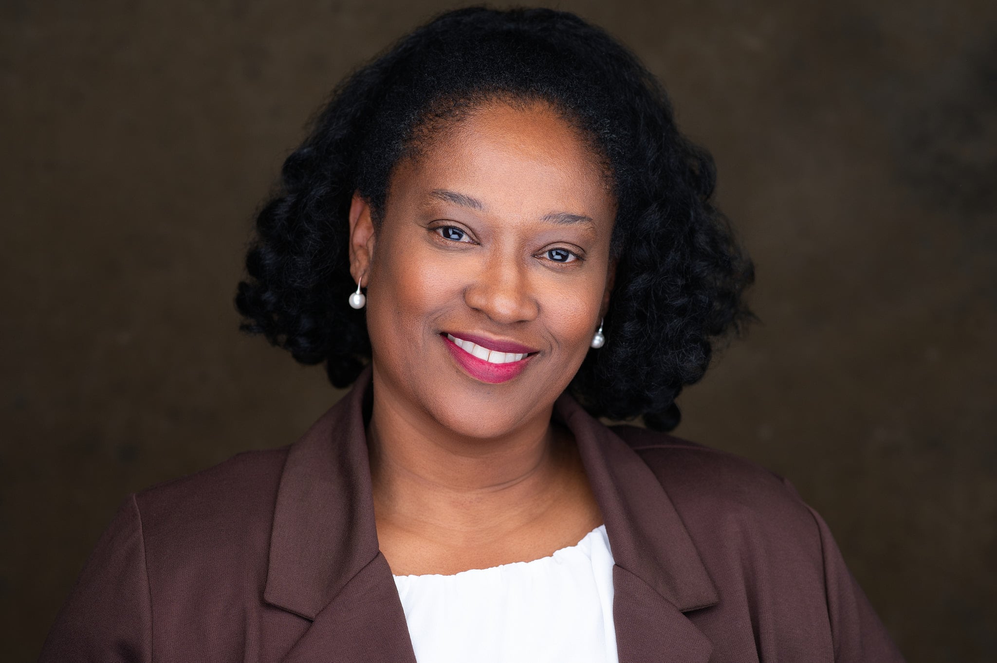 Professional entrepreneur headshot of woman in burgundy blazer with warm smile against soft muted olive background