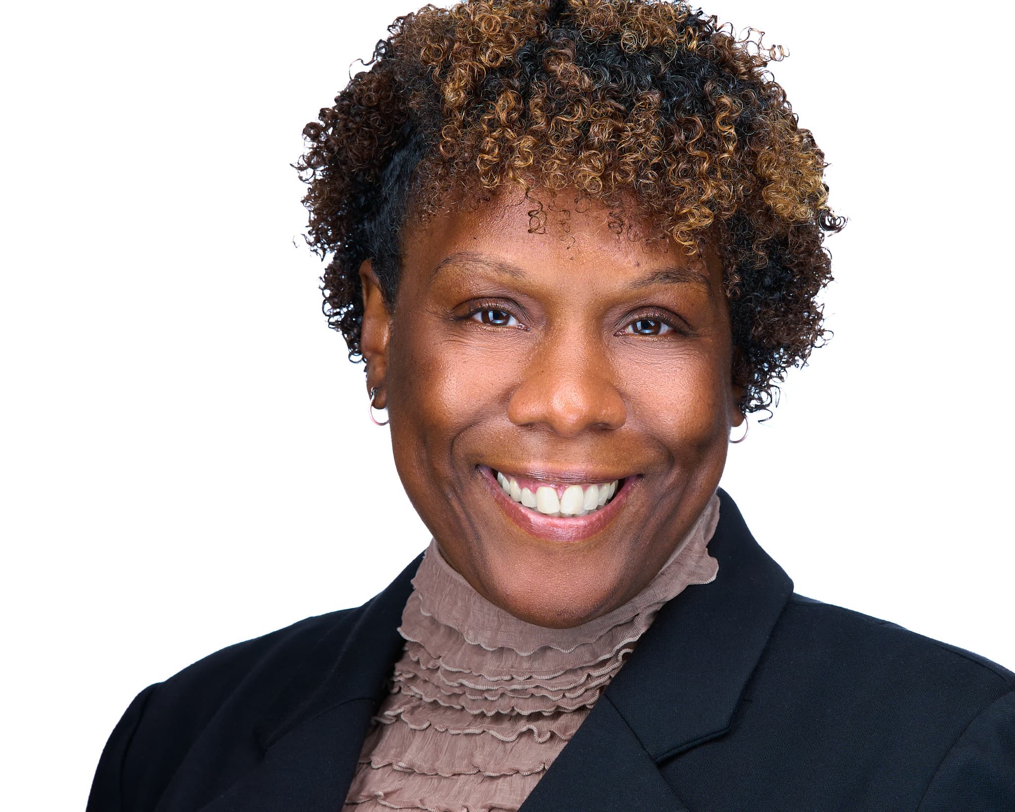 Professional headshot of a warm, approachable woman with short curled brown hair wearing a navy blazer and textured turtleneck in white studio setting