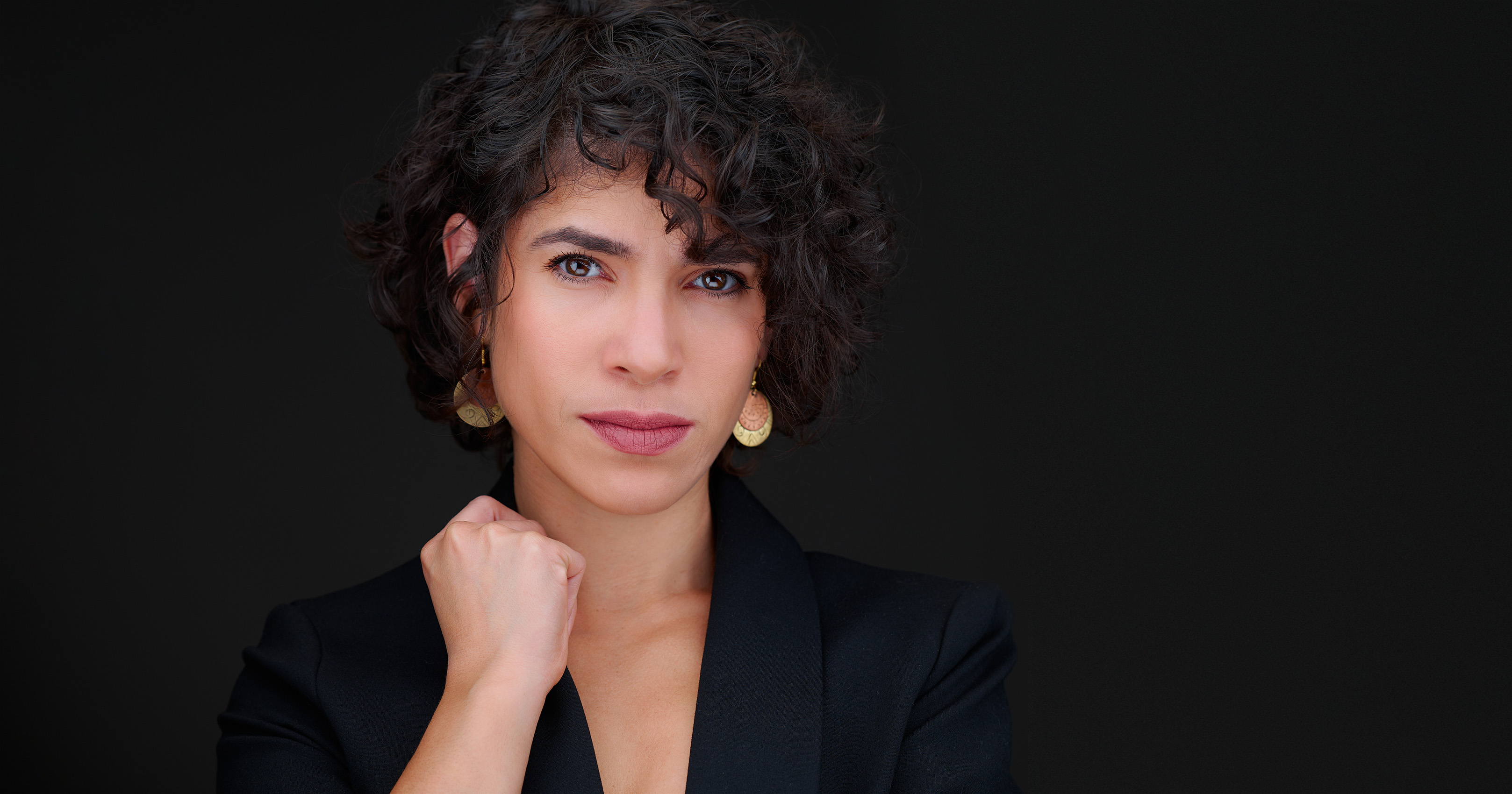 Professional headshot of a confident woman with short curly black hair and amber eyes wearing a black blazer against a clean black studio backdrop